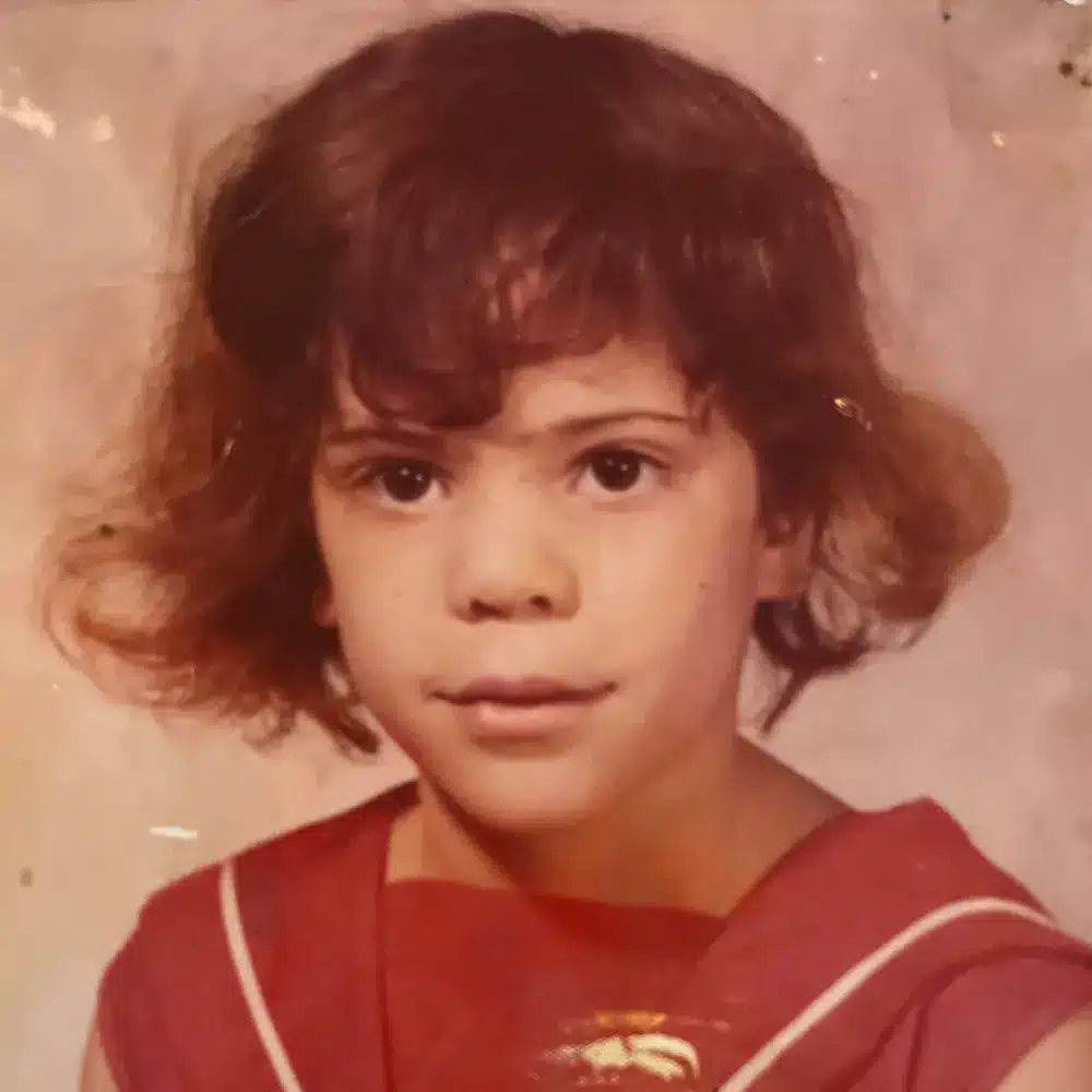 A young child with short, curly brown hair is wearing a red outfit with a white collar. The background is a plain, light color.