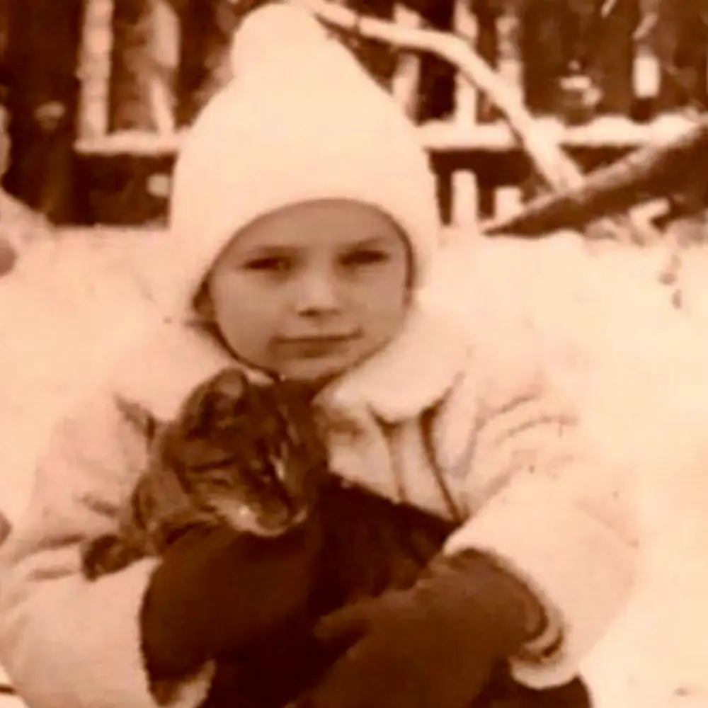 A child wearing a white coat and hat holds a cat in a snowy outdoor setting. The child is looking at the camera, and the scene is monochromatic, giving it a vintage appearance. Brown gloves are visible on the child's hands.
