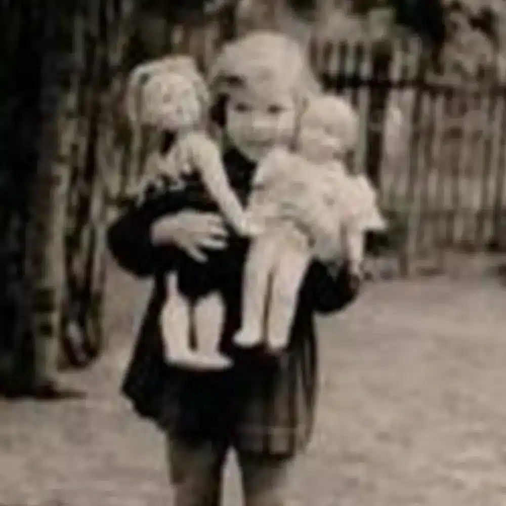 A black-and-white photo shows a young child standing outdoors, holding two dolls. The child is dressed in a dark checked coat. In the background, there is a tree and a blurred wooden fence.