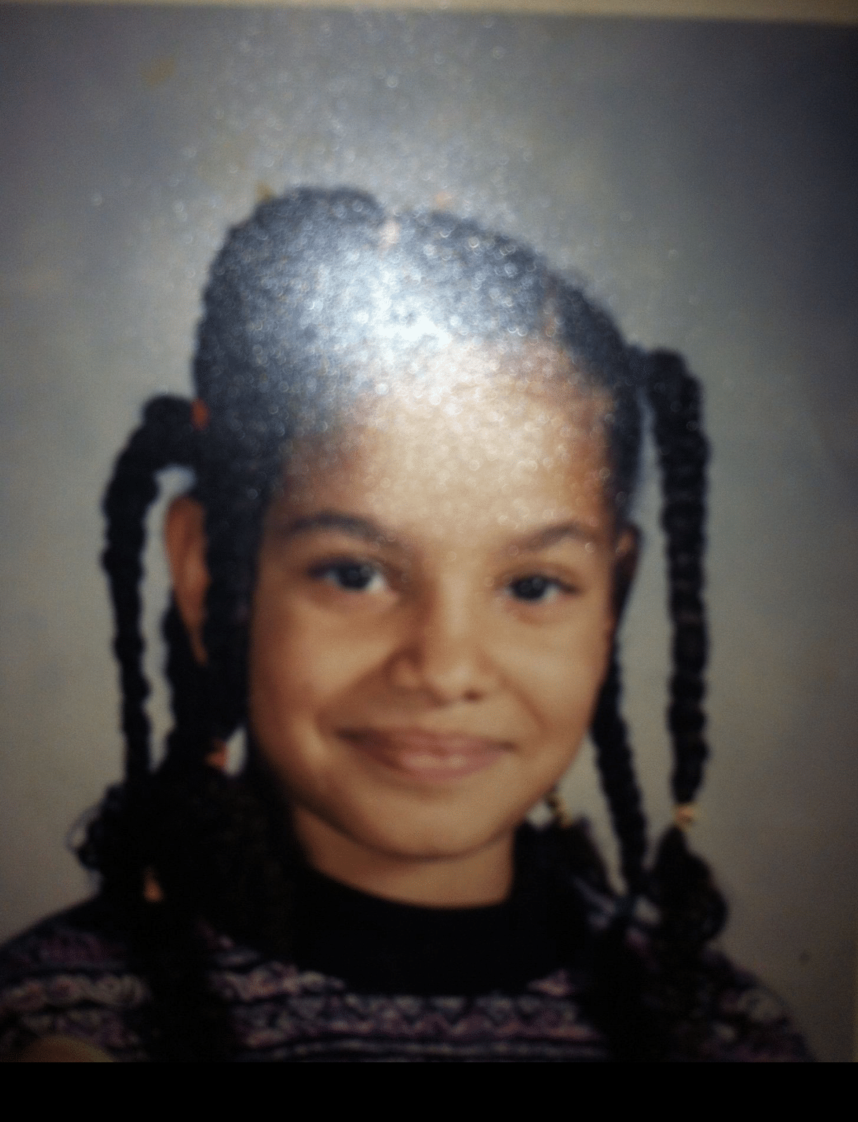A young girl with braided hair, wearing a patterned sweater, is smiling at the camera in a school portrait-style photo. There is a glare from a light reflecting on part of the image.